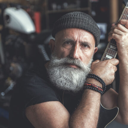 Confident bearded biker is siting in garage and holding black guitar. He glancing at camera with expectation. Portrait